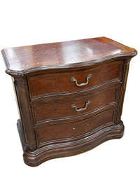 A dark brown wooden nightstand with two drawers and curved metal handles, photographed in what appears to be a furniture store or workshop.