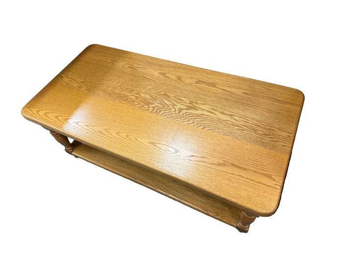 A rectangular wooden bench with rounded corners and a lower shelf, made from light oak wood with a glossy finish, shown in what appears to be a warehouse or furniture store.