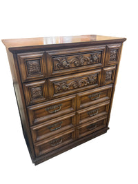 A dark brown wooden dresser with ornate carved drawer panels and metal handles, photographed in a warehouse or furniture showroom setting.