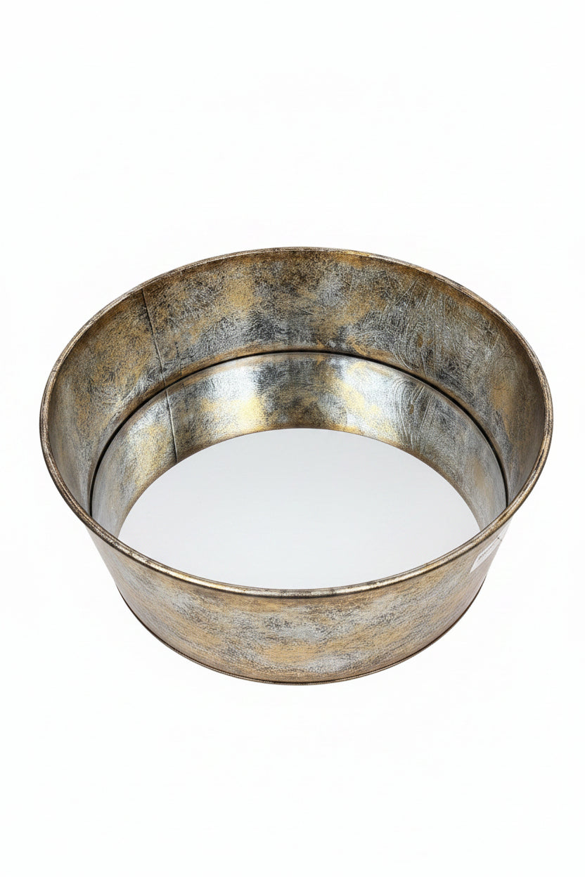 A round metal tray with a distressed, rustic silver and gold-toned finish, resting on a wooden table in front of a light wood cabinet.