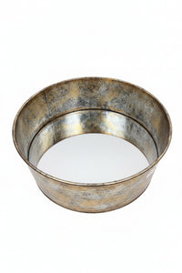 A round metal tray with a distressed, rustic silver and gold-toned finish, resting on a wooden table in front of a light wood cabinet.