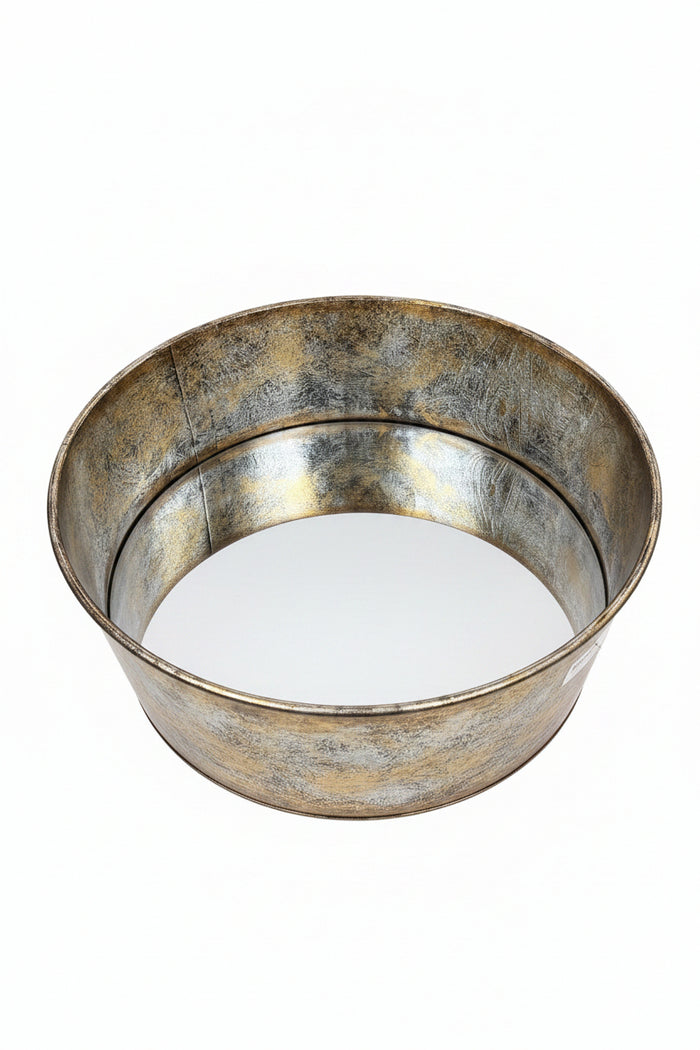 A round metal tray with a distressed, rustic silver and gold-toned finish, resting on a wooden table in front of a light wood cabinet.
