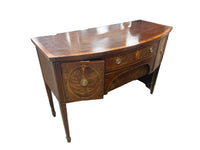 A curved wooden writing desk with a polished cherry wood finish, featuring a drawer front with a circular inlay motif and brass hardware, photographed in a warehouse setting.