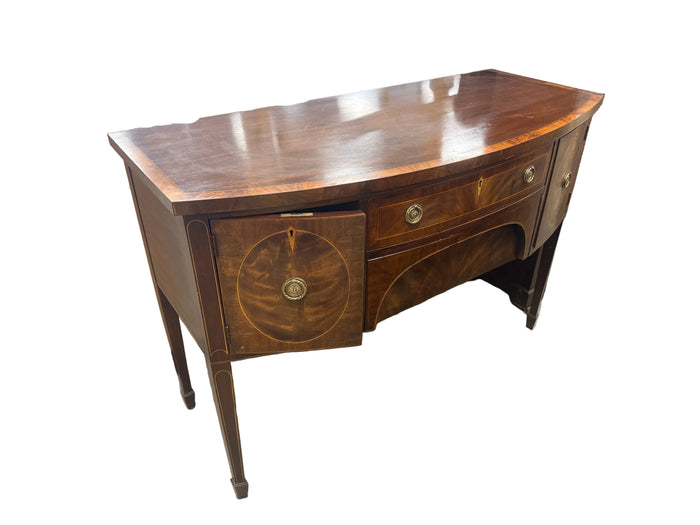 A curved wooden writing desk with a polished cherry wood finish, featuring a drawer front with a circular inlay motif and brass hardware, photographed in a warehouse setting.