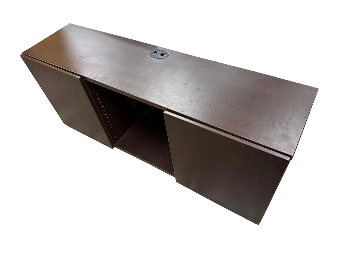A dark brown wooden office desk or console table with a circular cable outlet on top, photographed in a furniture store or warehouse setting with other furniture visible around it.