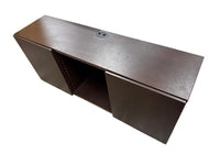 A dark brown wooden office desk or console table with a circular cable outlet on top, photographed in a furniture store or warehouse setting with other furniture visible around it.