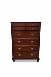 A dark reddish-brown wooden chest of drawers with six drawers and round metal pulls, photographed in a furniture store showroom.