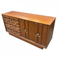 A mid-century wooden sideboard or credenza with a warm medium-brown finish, featuring a row of curved drawer pulls on the left and two cabinet doors with circular cutouts on the right, photographed in a furniture store.