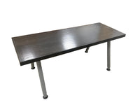 A rectangular desk with a dark gray wood-grain top and gray metal legs, shown in a warehouse or furniture store setting with other furniture and people partially visible in the background.