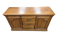 A medium brown wooden sideboard or credenza with three drawers in the center and two arched cabinet doors on the sides, shown in what appears to be a furniture store or warehouse.