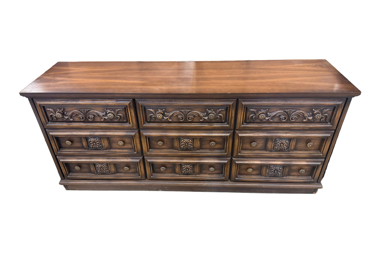 A dark brown wooden dresser with nine drawers and ornate carved drawer fronts, photographed in what appears to be a furniture store or workshop.
